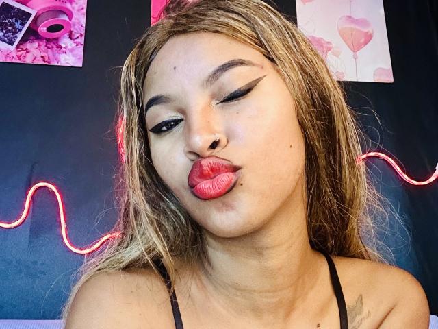 Plaissir69 with pouty red lips, winking, has blonde hair, black eyeliner, and a nose ring. Wearing a black strap, her tits are partially visible. Neon light wavy.