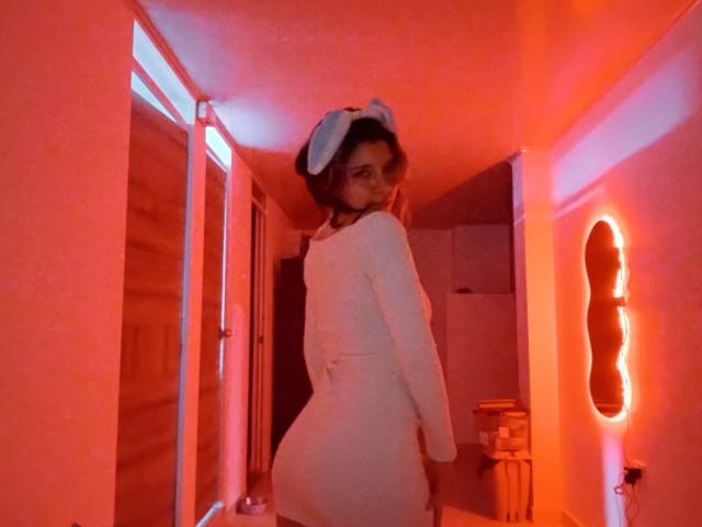 DarckVelvet is a sexy woman with a curvy ass, wearing a white bodysuit and blue bunny ears, standing in red-lit room, looking back with a sultry expression.
