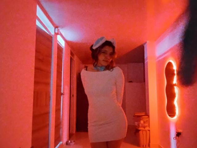 DarckVelvet stands in a room with red and blue lighting, wearing a white, long-sleeve, form-fitting dress and a blue headband, with hands behind her back, smiling.