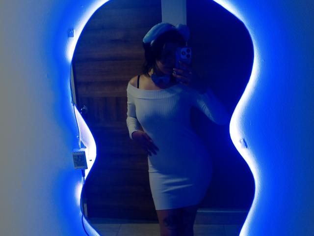 DarckVelvet stands in front of a mirror with a glowing blue outline, wearing a tight, off-shoulder white dress that accentuates her curvy figure. She has dark hair, a white hat, and holds a phone with a black and white case. The dress highlights her tattoos on her right thigh. The lighting is blue, creating a contrast with her white dress. The mirror's shape is irregular, adding a dynamic element to the composition.