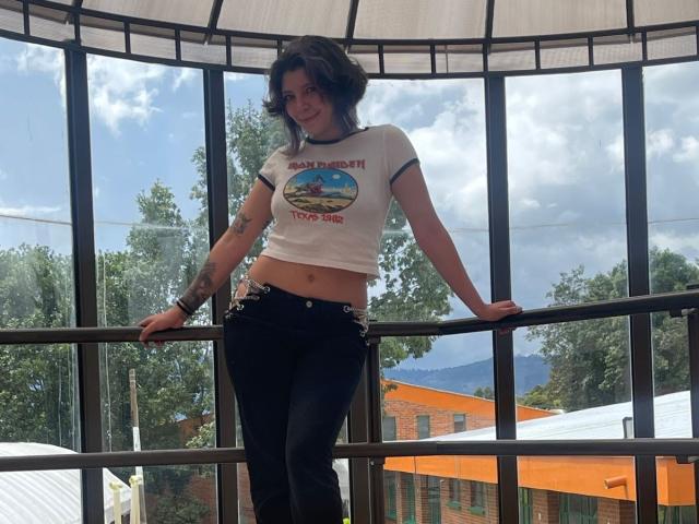 DarckVelvet with short dark hair, small tattoos, white crop top, black pants, leaning on glass railing, smiling, outside, sunny, trees, and buildings in the background.