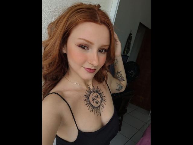 RuivaK with red hair, black tank top, sun tattoo on chest, tattooed arm, smiling, pink lipstick, white wall, and black fan in the background.