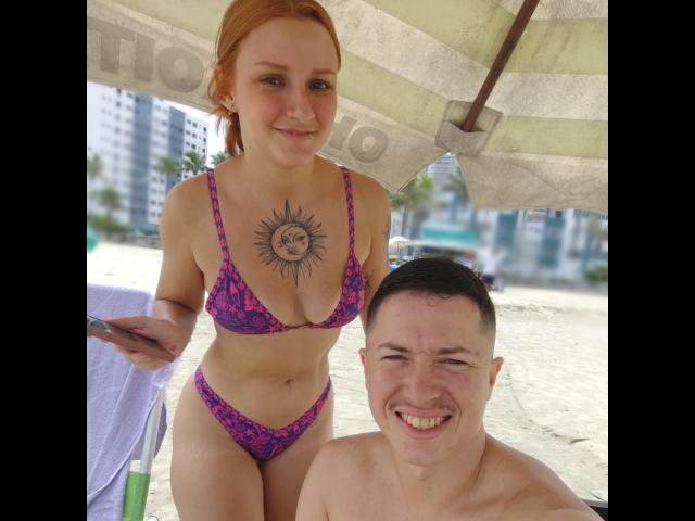 RuivaK with red hair, sun tattoo on chest, pink and purple bikini, stands next to a smiling, shirtless guy with short dark hair, under a beach umbrella.