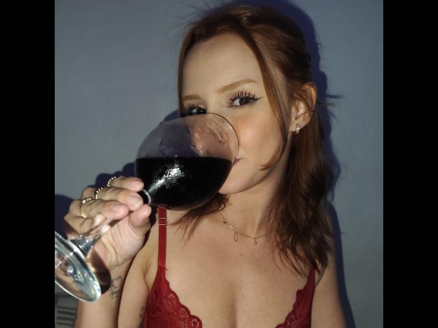 RuivaK with red lace bra, drinking dark wine from a glass, her lips on the rim, showing off her tits, brown hair, and eyeliner, wearing a necklace, against a blue wall.