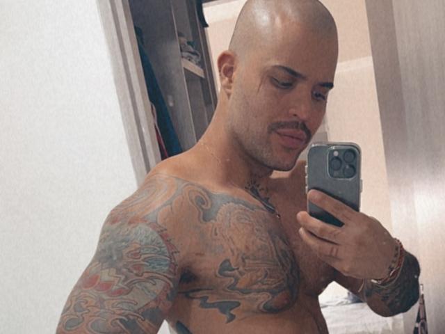 MedellynDark est un homme chauve, tatoué, torse nu, avec une moustache, tenant un téléphone, prenant un selfie, et montrant ses tatouages colorés sur ses bras et sa poitrine..