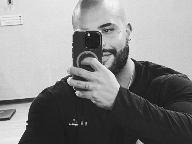 MedellynDark, bald with a beard, holding a phone, sticking out his tongue, wearing a black long-sleeve shirt, in a black-and-white selfie.