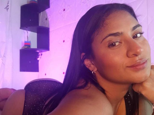 DiannaRosse, met lang zwart haar, bruine ogen en een lichte glimlach, ligt neer, waarbij haar blote schouders en een kleine, glanzende neus zichtbaar zijn, met een paars licht op de achtergrond..
