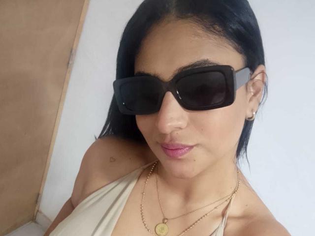DiannaRosse has black hair, wearing big black sunglasses, pink lipstick, a gold necklace, and a white top, showing a small heart tattoo on her shoulder.