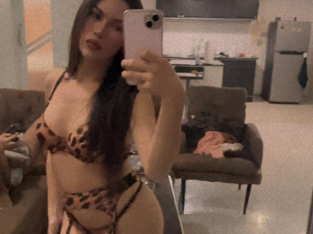 KissmePaula, avec de longs cheveux noirs, portant un ensemble de soutien-gorge et jarretière à motifs léopard, se prend en photo devant un miroir, montrant ses seins et ses fesses, dans une pièce peu éclairée..