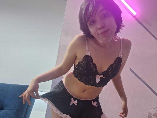 ElodiePerrine with short black hair, wearing a black lace bra with pink bows and a matching black mini skirt with pink bows, leaning forward, showing her small tits and flat ass.