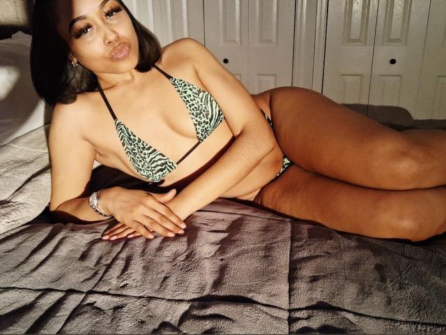 ChanelHope lying on a bed in a green and black zebra-pattern bikini, showing her tits and ass, with a pouty face, dark hair, and a bracelet on her wrist.
