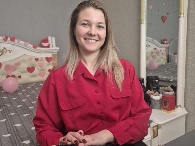 Anshante, con i capelli biondi, sorridente, indossa una camicia rossa con bottoni, ha un seno piccolo ed è seduta in una camera da letto con un letto a motivi di cuori bianchi e rosa..