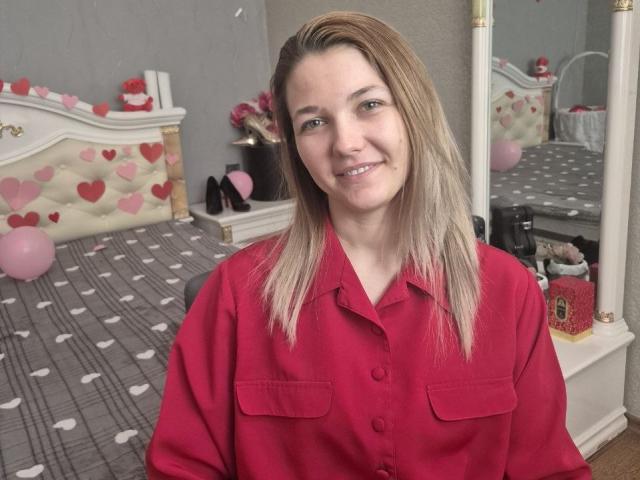 Anshante with blonde hair, wearing a red button-up shirt, smiling in a bedroom with pink heart-patterned bed and mirror. Her tits are average, her ass is not visible.
