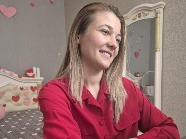 Anshante a la peau claire et les cheveux blonds et lisses, et elle porte une chemise rouge à boutons. Elle sourit légèrement et regarde vers la droite. Ses sourcils sont bien dessinés, et elle a un maquillage subtil pour les yeux, avec un trait fin. L'arrière-plan comprend un miroir blanc orné avec des détails dorés, et un lit avec un motif à carreaux gris et blanc, décoré de cœurs roses. L'image a un style réaliste et décontracté..