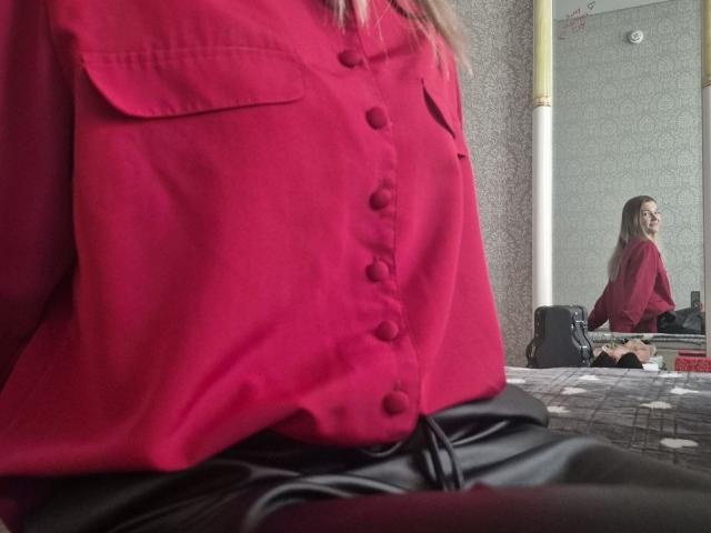 Anshante trägt ein rotes Hemd mit Knopfleiste, schwarze Lederhosen. Im Spiegelbild ist sie zu sehen, mit dem Rücken zur Kamera, langes blondes Haar, weiße Wand, gelöste Zigarette im Hintergrund..