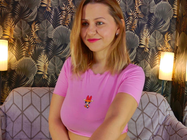 NymphoArri with blonde hair, pink crop top, and small tits, smiling, wearing pink lipstick, standing in front of a patterned wall, lit by two lamps on either side.
