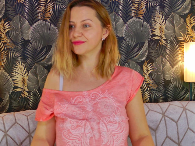NymphoArri with blonde hair, pink shirt, white bra strap, and pink lipstick, sitting on a gray patterned couch, smiling softly. Her tits are visible under the shirt.