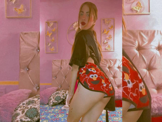 Sharktitiare with red anime-print booty shorts, black t-shirt, and pink lipstick, looking over shoulder, showing ass, in pink room, tufted bed, and floral pillows.