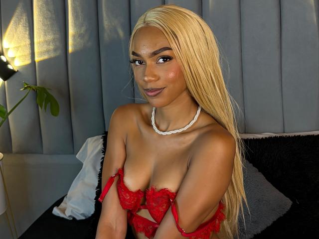 AylaCallowey with long blonde hair, wearing red lace bra, exposing her small tits, white necklace, sitting on bed, smiling, dark skin, black pillow, gray curtain in background.