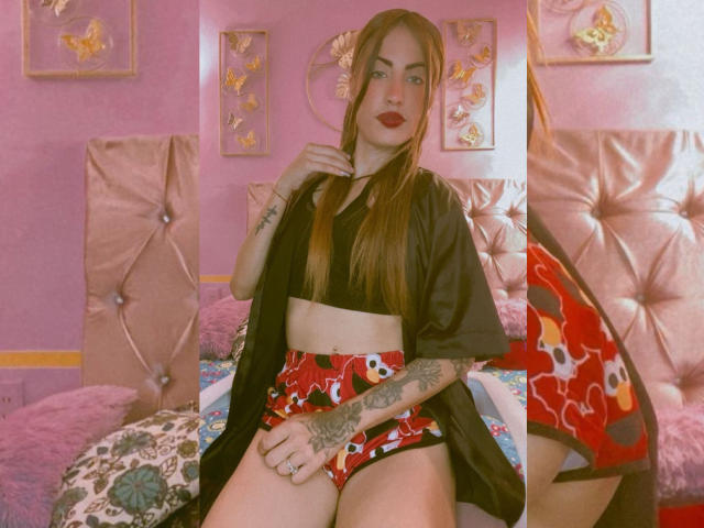 Sharktitiare with long red hair, black crop top, red patterned panties, black open shirt, tattoos, sitting on bed, pink wall, tufted headboard, and floral wall art.
