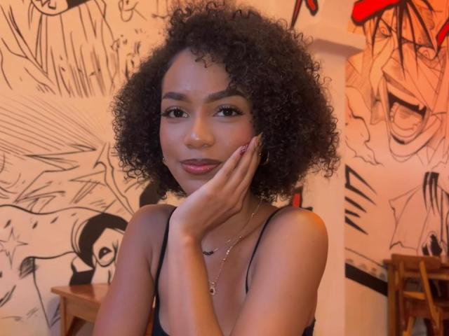 A photograph of AylaCallowey with curly black hair, brown skin, and a cute face, touching her cheek with her hand, wearing a black tank top, and a necklace.