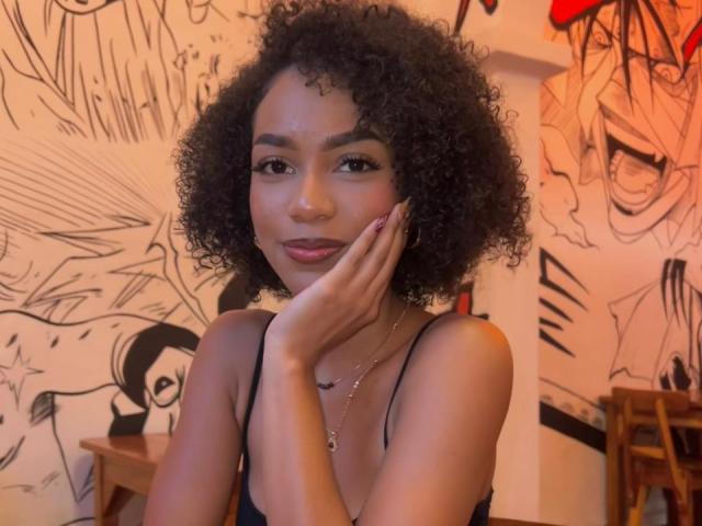 A photograph of AylaCallowey with curly black hair, light brown skin, and a cute smile, touching her cheek with her hand, wearing a black spaghetti strap top.