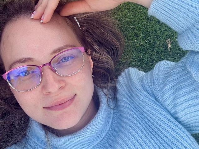 AmbeerWood with pink glasses, blue turtleneck sweater, lying on grass, smiling, hand in brown hair, light skin, freckles, cute, relaxed, blue eyes, pinkish nose, pink nails.
