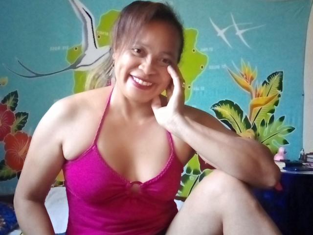 Rahel69, smiling, brown-haired, in pink halter top, showing small tits, sitting with knee up, hand on cheek, pink lipstick, blue-green background with floral and bird patterns.