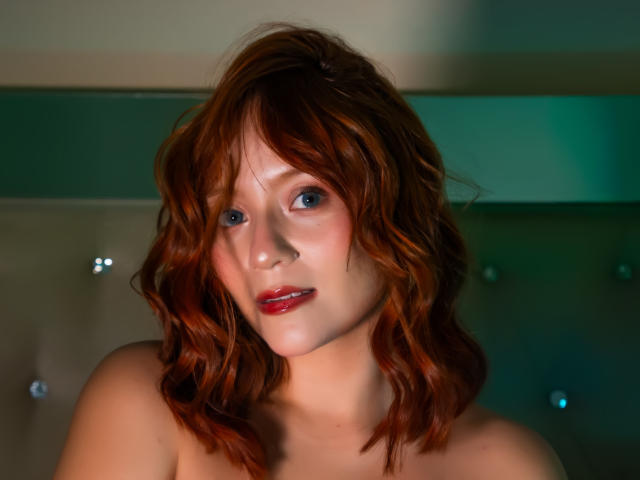 EmilyKlein has red, wavy hair, blue eyes, and red lips. She's topless, showing her bare shoulders and soft skin. Her cute, sexy face is slightly tilted.