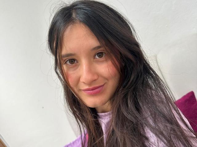 KimyRosse has long, messy black hair, brown eyes, and a pinkish nose with a slight smile, wearing a purple top, showing soft, natural-looking tits in the photo.