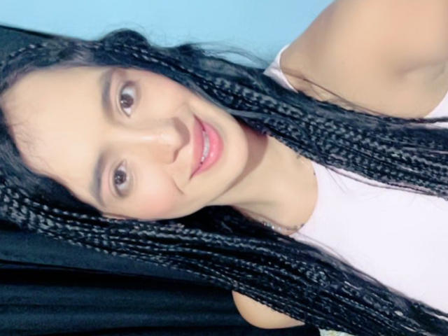 HeraDiosa a de longues tresses noires, des yeux marron et un sourire rose, elle porte un débardeur blanc, avec un fond bleu clair et un rideau noir..