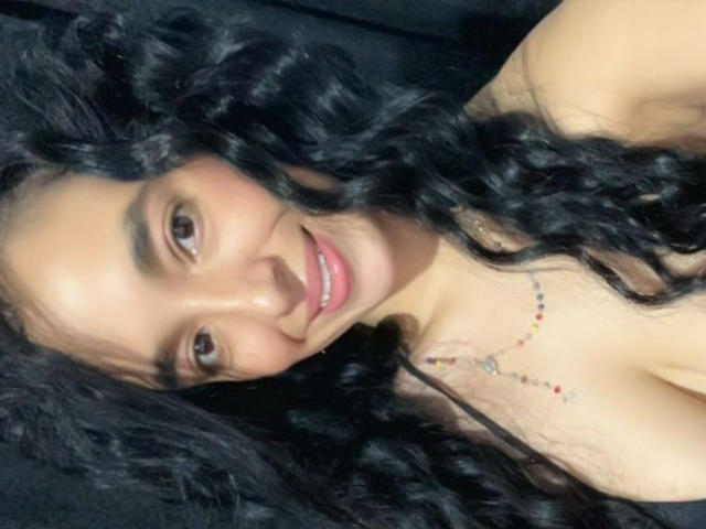 HeraDiosa with curly black hair, brown eyes, and a smile, showing her bare shoulders and a necklace with small red and blue beads. Her tits are partially visible.