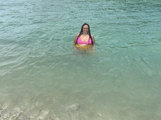 HeraDiosa with large, pink bikini top, water up to her chest, smiling, greenish-blue water, brown hair, yellow inflatable, standing in shallow, clear, rocky beach.