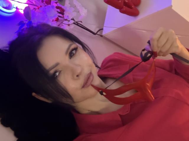 MollyBrunett with black hair, wearing a red shirt, holding a red dildo with her lips. Her eyes are half-closed, and she's lying on a pink surface.