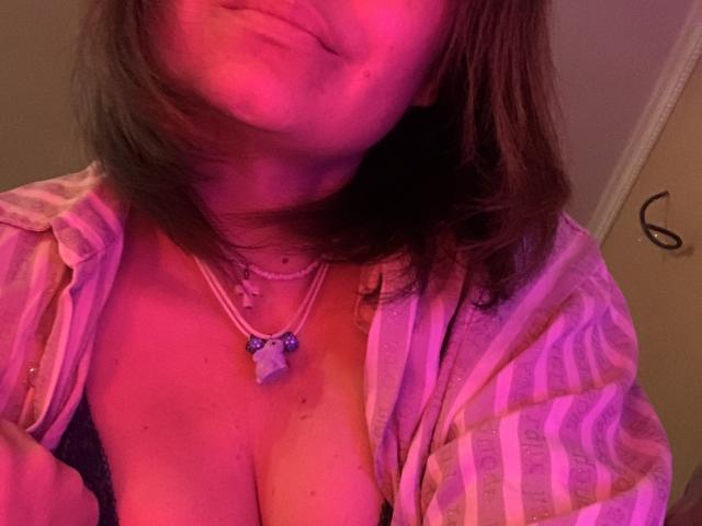 ElizavetaCream's face and chest in pinkish-purple light, wearing an open striped shirt, necklace with blue beads, exposing her large boobs, slightly pouty lips, brown shoulder-length hair.