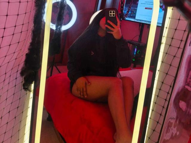 Zarafadi in un selfie scattato allo specchio, indossa una felpa nera, è seduta su un letto rosso, con le gambe incrociate, tiene un telefono in mano, ha i capelli lunghi, c'è una luce ad anello, degli specchi a LED e uno schermo del computer..