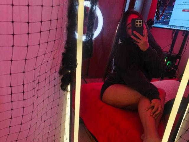 Zarafadi tirando uma selfie no espelho, vestindo um moletom escuro, shorts pretos, uma faixa de cabelo vermelha, sentada em uma cama vermelha, com uma rede à esquerda, segurando um celular na mão..