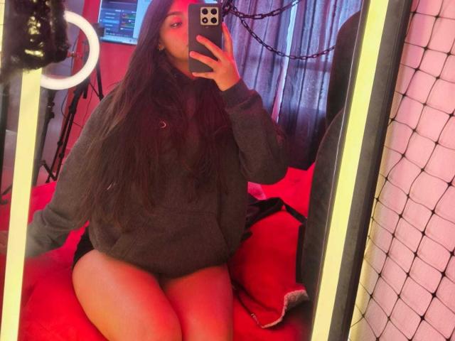 Zarafadi, con lunghi capelli scuri, indossa un maglione grigio e biancheria intima nera, seduta su un letto rosso, sta scattando un selfie allo specchio con un iPhone, mostrando il suo sedere..