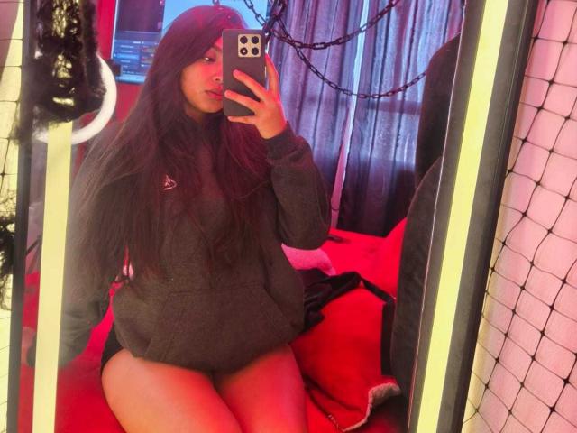 Zarafadi, met lang donker haar, draagt een grijze hoodie en zit op een rood bed. Ze houdt een telefoon vast en toont haar blote billen in een spiegelzelfportret..