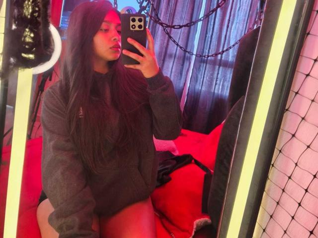 Zarafadi, con cabello largo y oscuro, vistiendo un suéter gris, tomando una selfie con un espejo, mostrando muslos desnudos, iluminación roja, y sosteniendo un teléfono negro, en una habitación con poca luz..