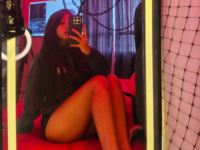 Zarafadi, con lunghi capelli neri, indossa una maglietta nera a maniche lunghe e biancheria intima nera, seduta su una sedia rossa, sta scattando un selfie allo specchio, mostrando il sedere e le gambe scoperte..