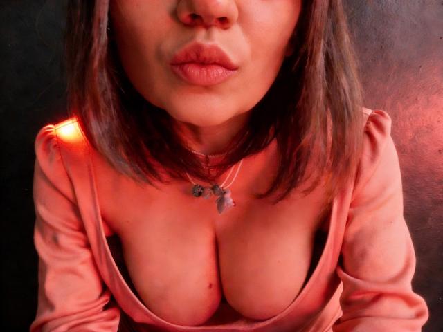 ElizavetaCream has puffy pink lips, shoulder-length brown hair, and a pink top revealing her large tits. She wears a necklace with small star-shaped pendants.