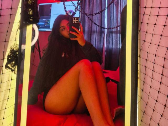 Zarafadi, con cabello largo y oscuro, vestida con un suéter negro, sentada en una cama roja, con las piernas cruzadas, mostrando su trasero desnudo, tomando una selfie con el espejo usando un teléfono..