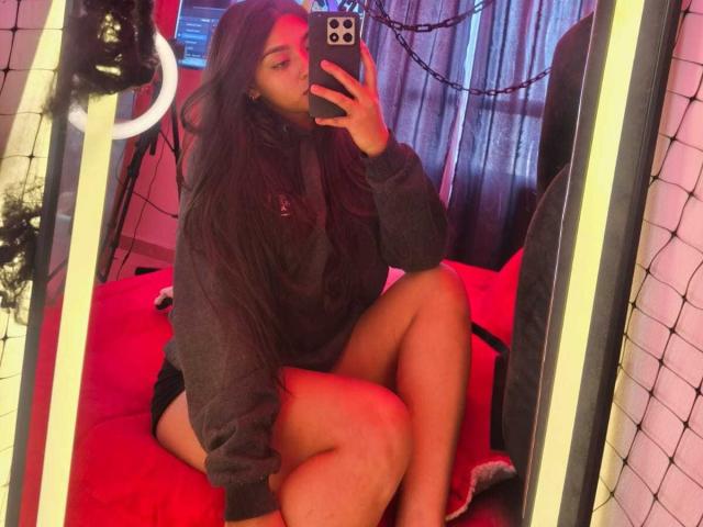 Zarafadi, con lunghi capelli scuri, indossa una felpa grigia e biancheria intima nera, seduta su un letto rosso, sta scattando un selfie allo specchio con il telefono, mostrando le gambe e i glutei..