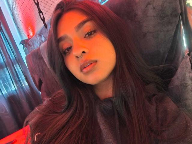 Zarafadi heeft lang donker haar, een neusring en volle, roze lippen. Ze draagt een grijze hoodie en kijkt naar de camera met een lichtjes open mond. Rood licht accentueert haar gezicht..