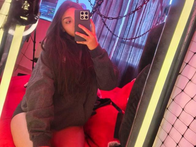Zarafadi, con cabello largo y oscuro, vestida con una sudadera gris, tomando una selfie frente al espejo mostrando su trasero desnudo, sosteniendo un teléfono con una luz de anillo en el fondo..
