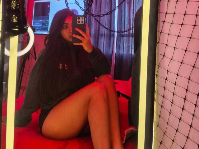 Zarafadi met lang donker haar, een zwart shirt en zwarte korte broek, zittend op een rood bed, met een telefoon in haar hand, spiegelbeeld, benen gekruist, een zwart net aan de rechterkant, een ringlamp aan de linkerkant..