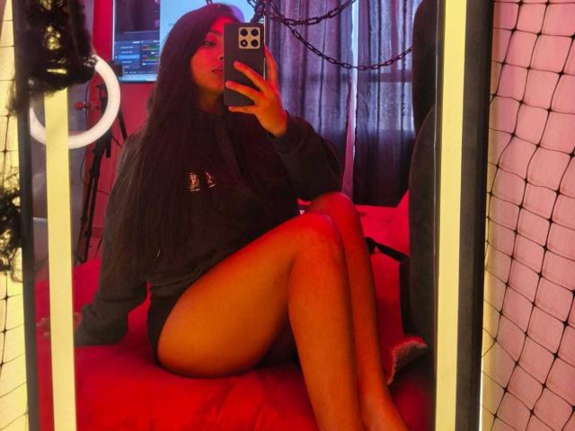Zarafadi mit langem, schwarzem Haar, die einen dunklen Pullover trägt und auf einem roten Bett sitzt, die Beine gekreuzt hat und ein Selfie mit ihrem iPhone macht, wobei ihr nackter Hintern zu sehen ist..