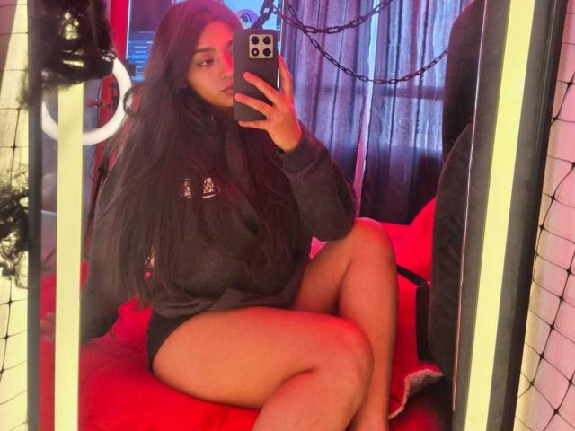 Zarafadi, com cabelos longos e pretos, vestindo um moletom escuro e shorts pretos, está sentada em uma cadeira vermelha, tirando uma selfie no espelho com um celular, mostrando suas coxas nuas..