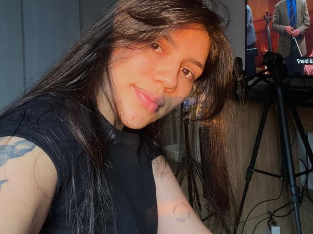 SophiaMarx with long brown hair, black shirt, tattoo on left arm, pink lips, smiling, television on tripod in background, visible on right.